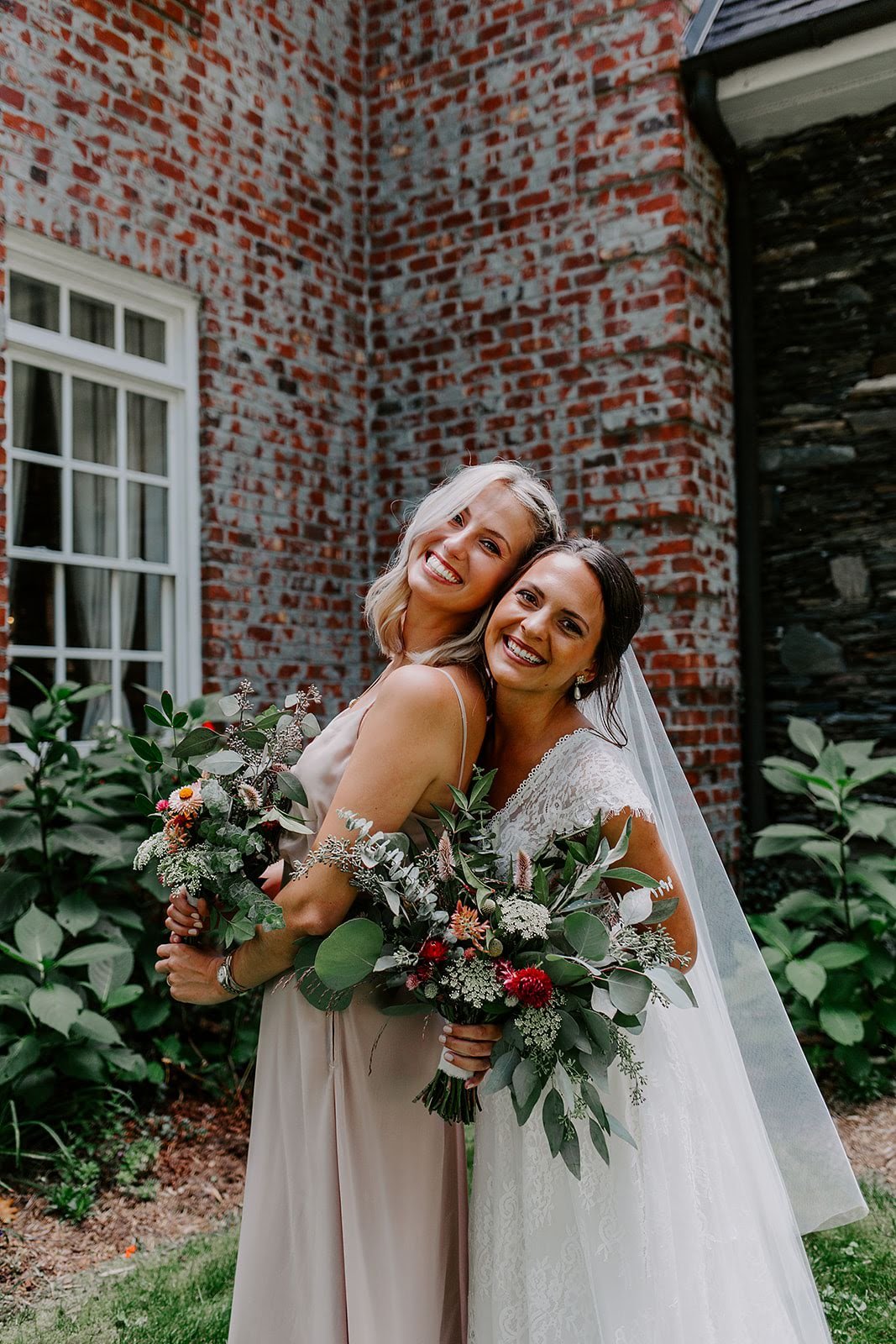 Bride and bridesmaid smiling together with bouquets beside a brick estate wedding venue.