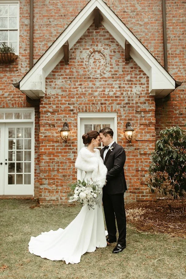 Bride and groom kissing in front of a brick venue under a peaked gable, bride wearing a fur wrap.