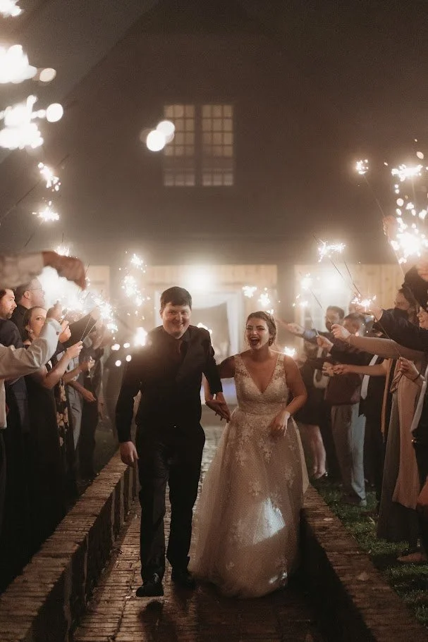 Newlyweds walking hand in hand through a sparkler exit surrounded by cheering wedding guests at night.
