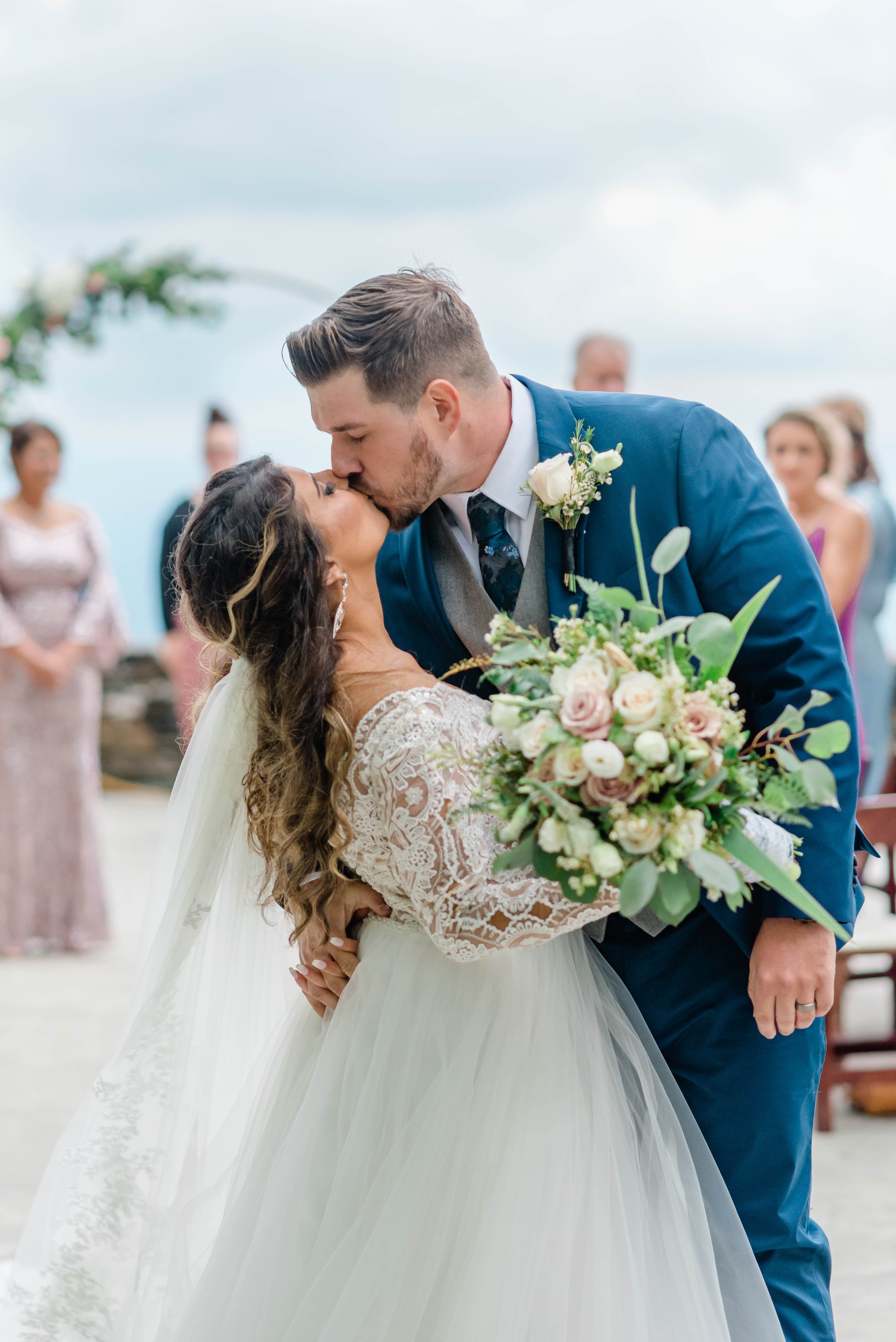 Bride and groom kissing during their outdoor wedding ceremony with a blush and greenery bouquet.