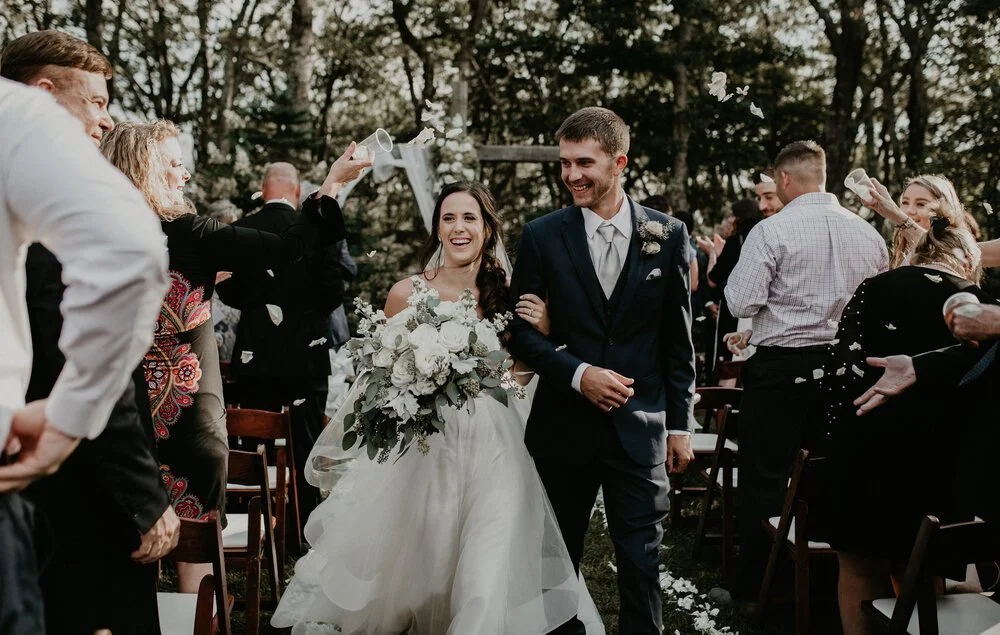 A joyful bride and groom walk down the aisle after their outdoor wedding ceremony as guests shower them with white petals.