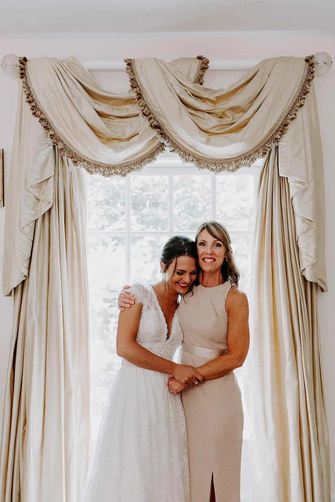 Bride and mother standing together for a portrait by a bright window before the ceremony.