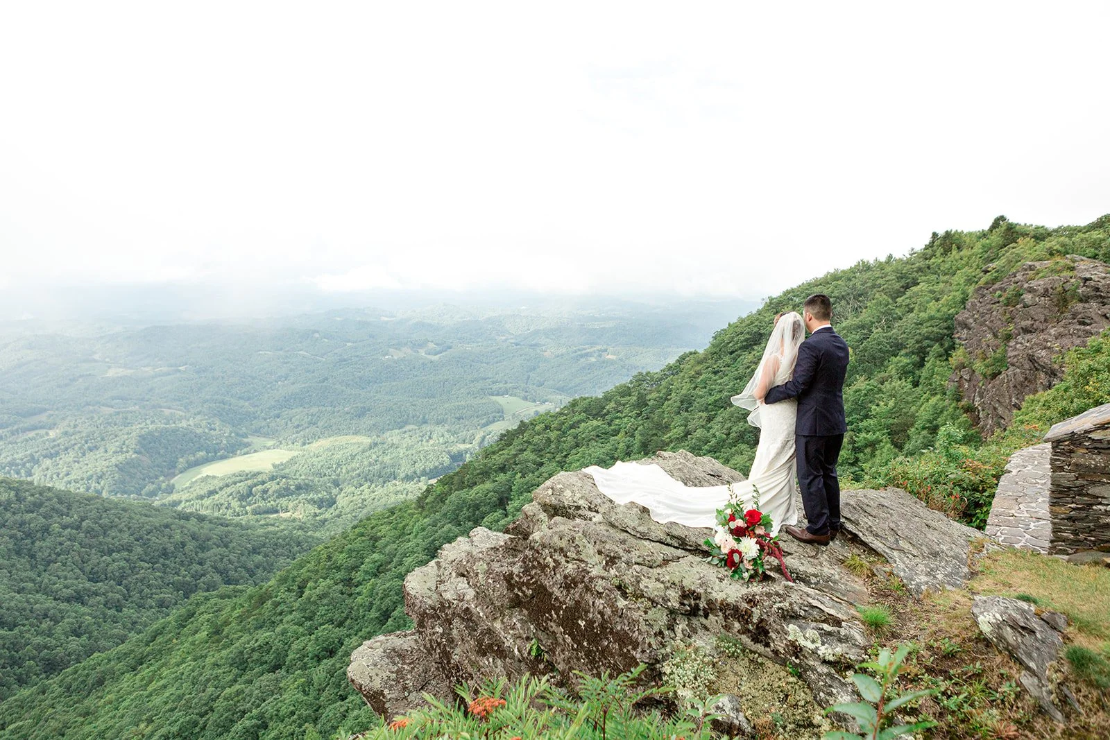Bride and groom standing on a mountain cliff with the bride’s long veil and bouquet overlooking the valley.