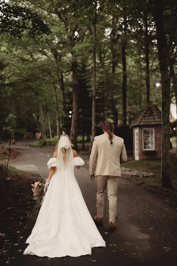 A bride in a white gown and groom walk away on a paved path through a lush green forest, towards a small rustic building.