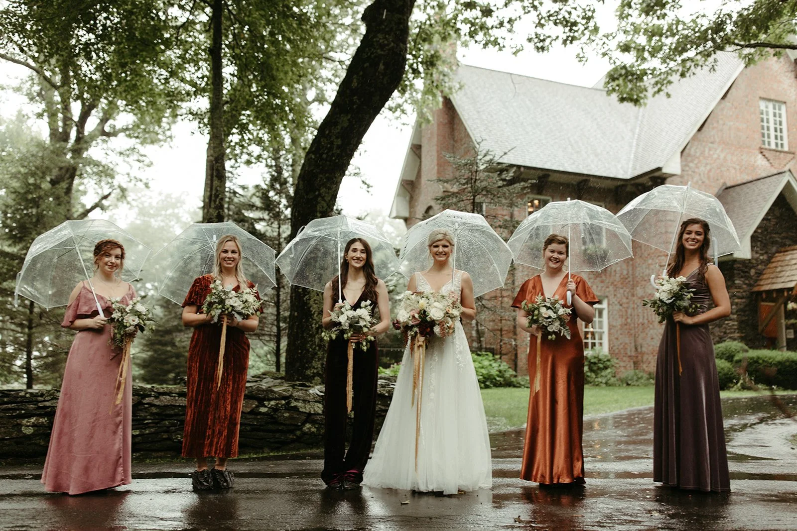 Bride and bridesmaids holding clear umbrellas in the rain outside a brick wedding venue