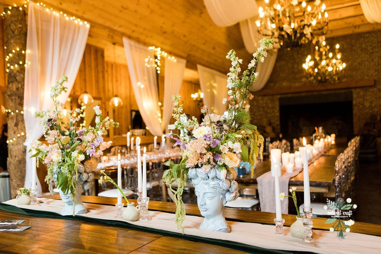 Rustic barn reception decor with floral centerpieces, candles, and white draping under chandeliers