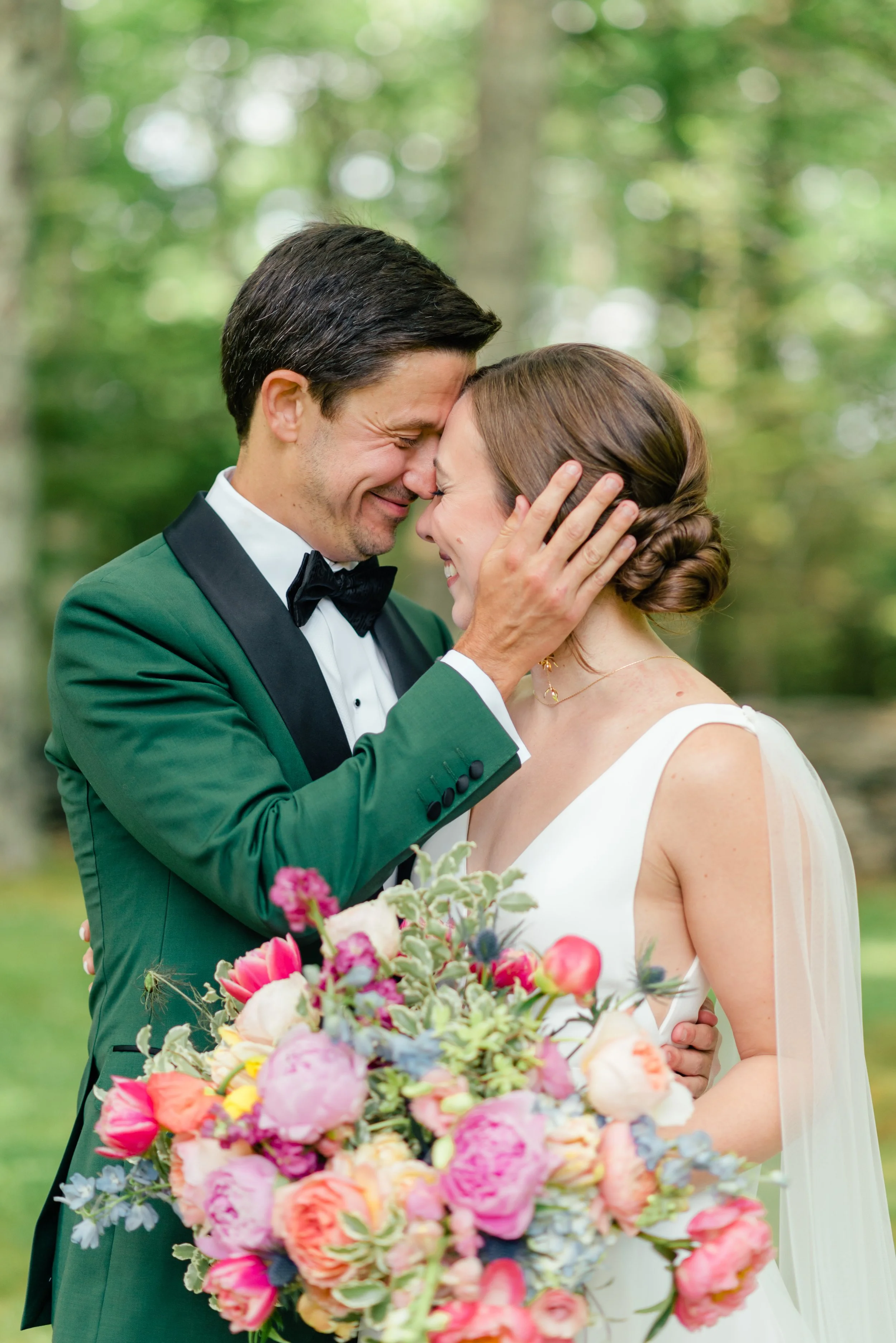 Bride and groom touch foreheads and smile in a wooded setting, holding a colorful wedding bouquet.