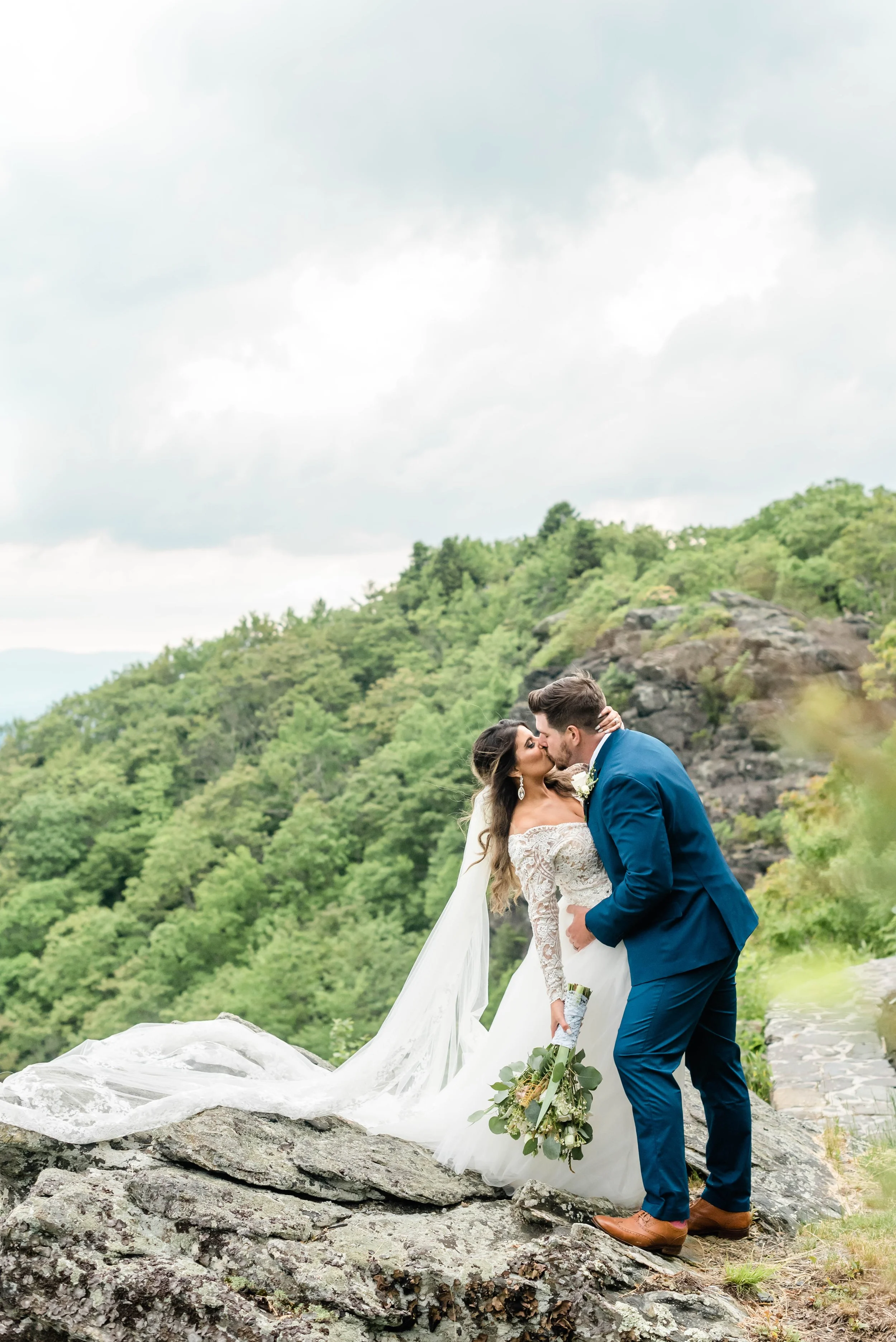 Bride and groom kissing during mountain portraits, groom in a blue suit and bride holding a bouquet