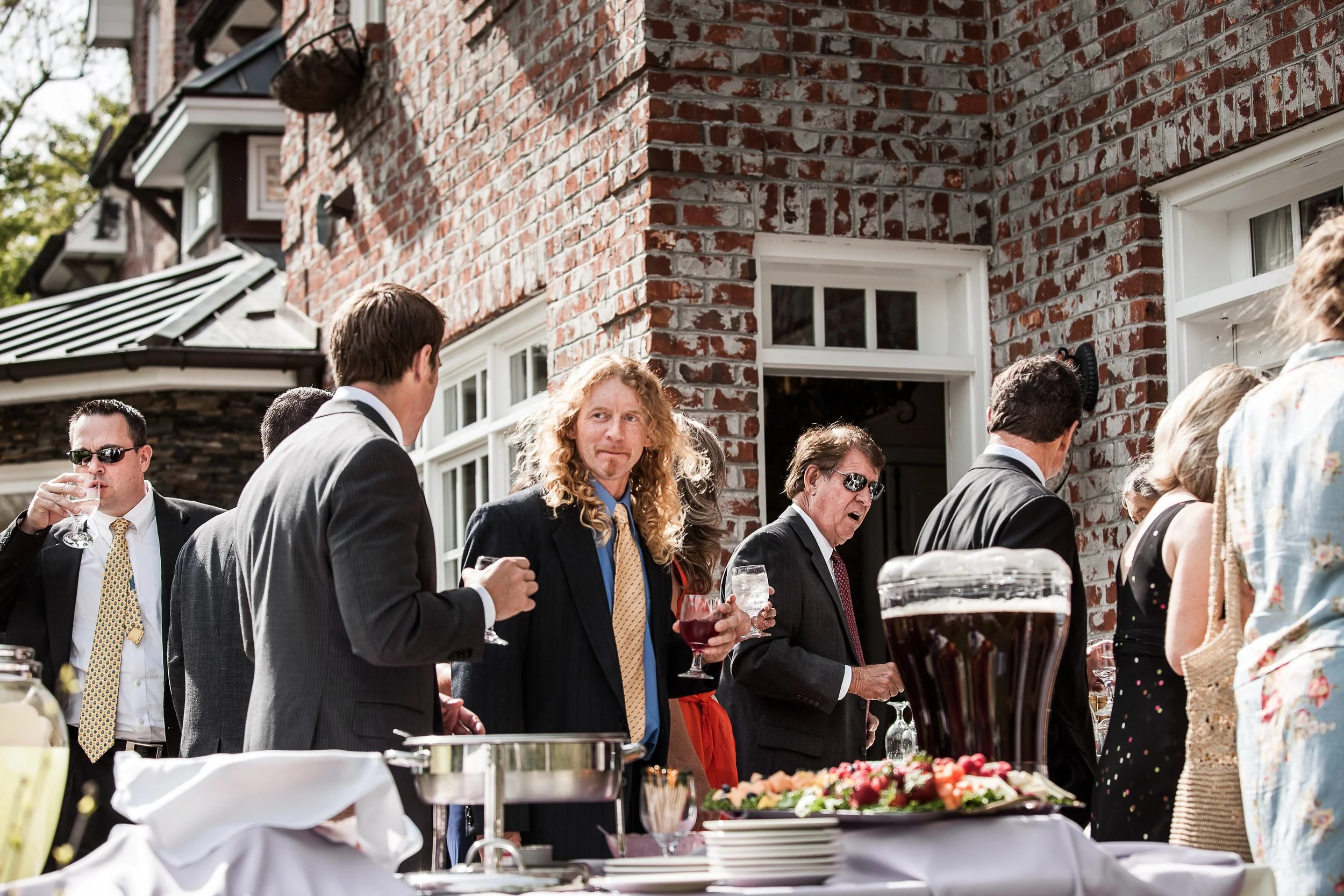 Wedding guests enjoying cocktail hour outside a historic brick estate with drinks, appetizers, and buffet tables.