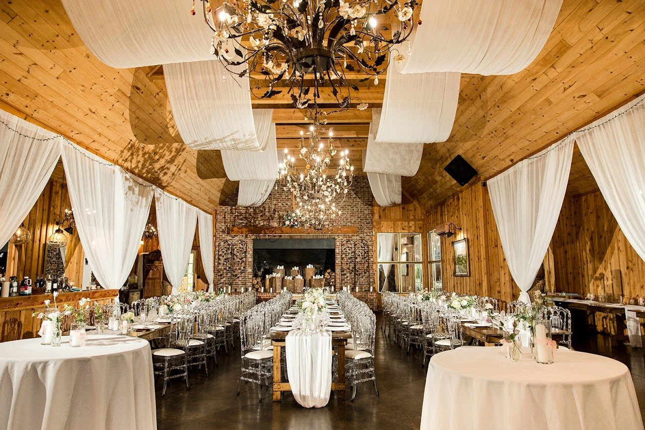 A rustic wooden barn reception hall decorated with white draping, crystal chandeliers, and long banquet tables.