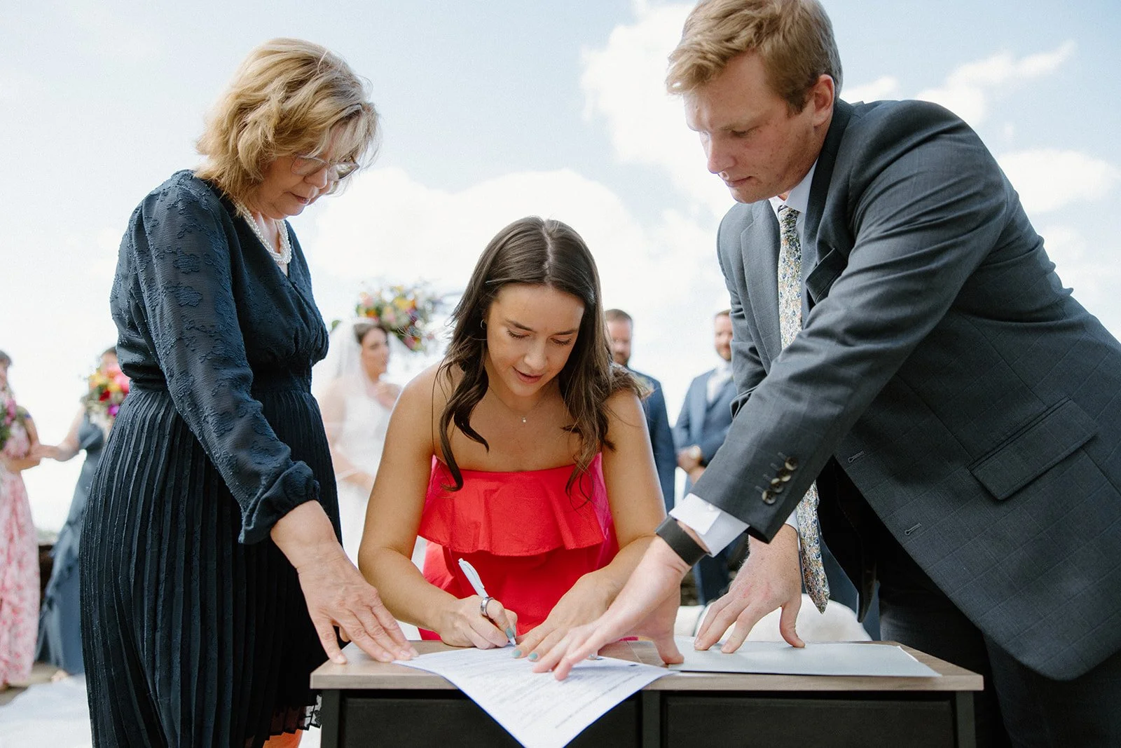 Couple signing the wedding license outdoors with family members assisting at the table.