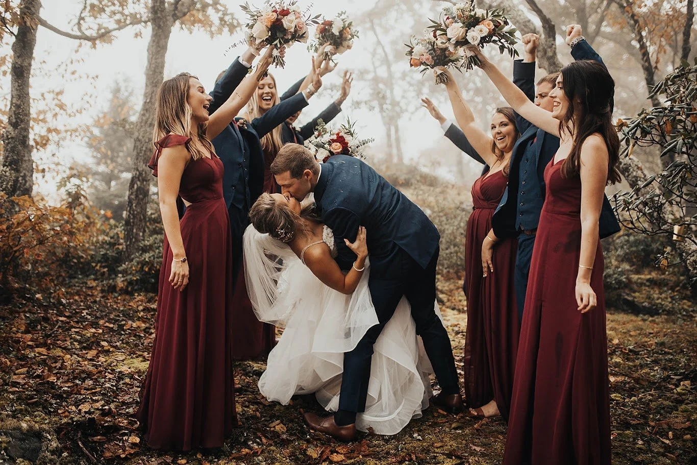 Wedding party cheering as the groom dips the bride for a kiss in the woods.