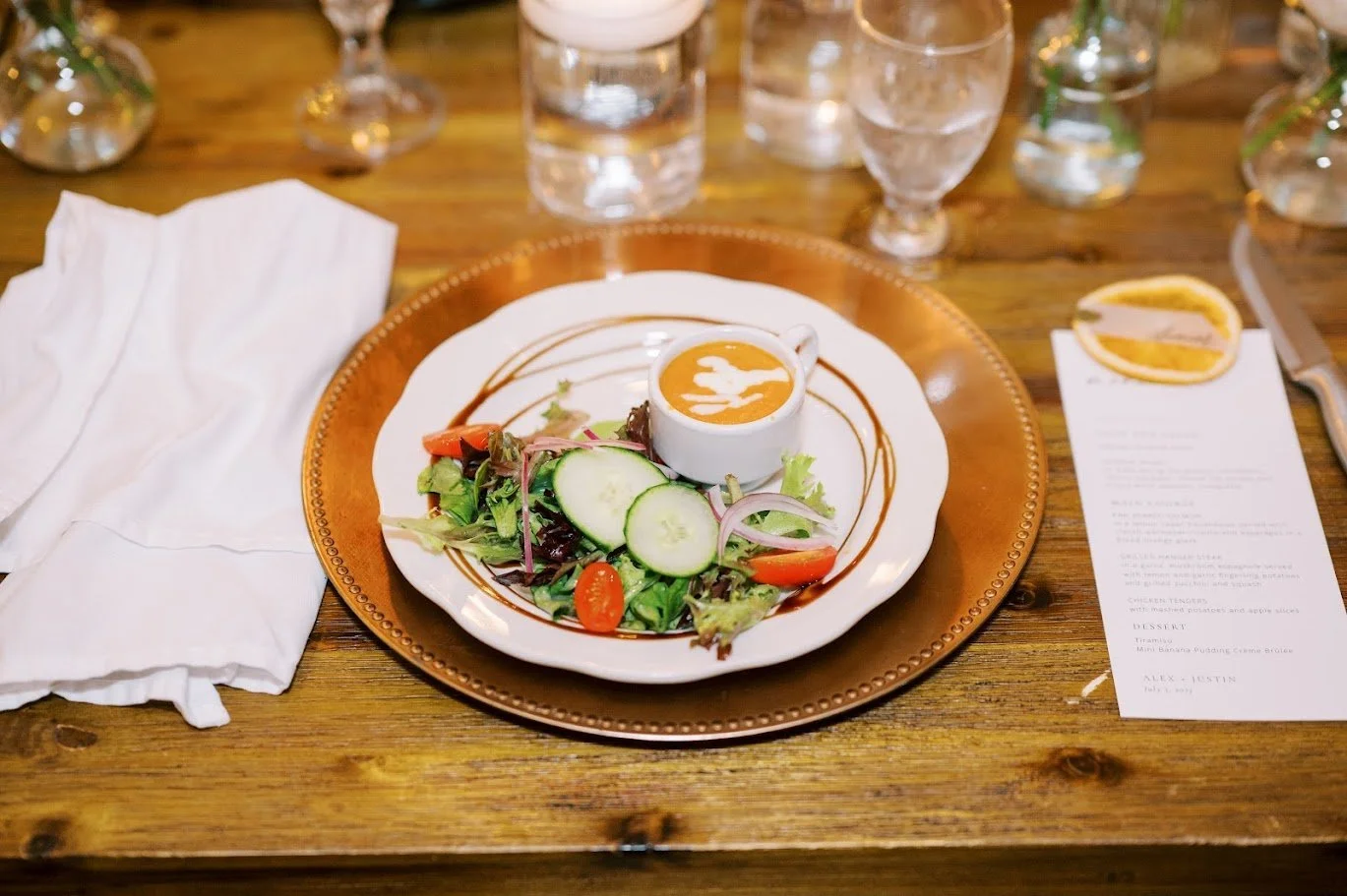 An elegant place setting features a salad with greens, tomatoes, cucumbers, and a small white bowl of soup, on a gold charger, with water glasses and a menu card.