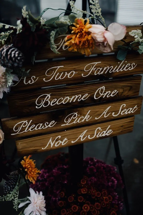 Wood wedding sign reading “As two families become one, please pick a seat not a side” decorated with flowers.