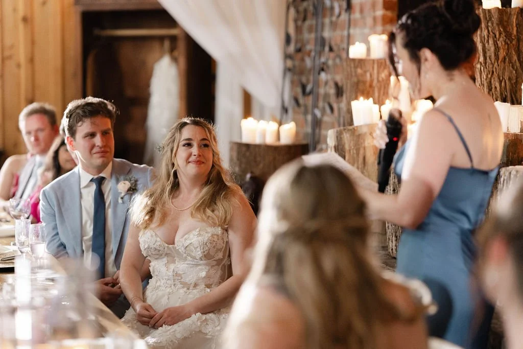 Bridesmaid giving a speech while the bride and groom listen at a candlelit rustic wedding reception.