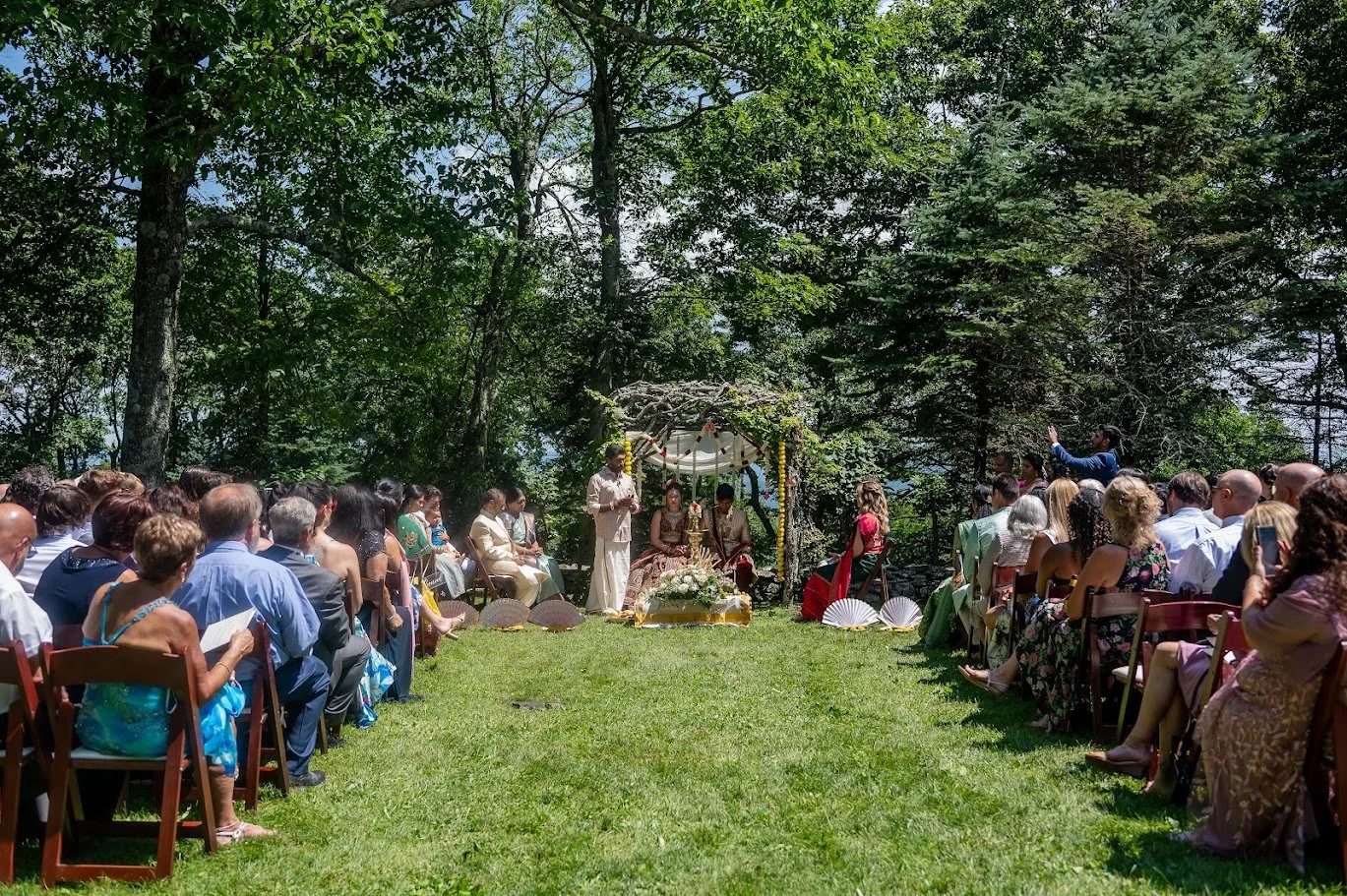 Guests seated on a grassy aisle facing a floral-draped canopy, with trees framing the outdoor ceremony.