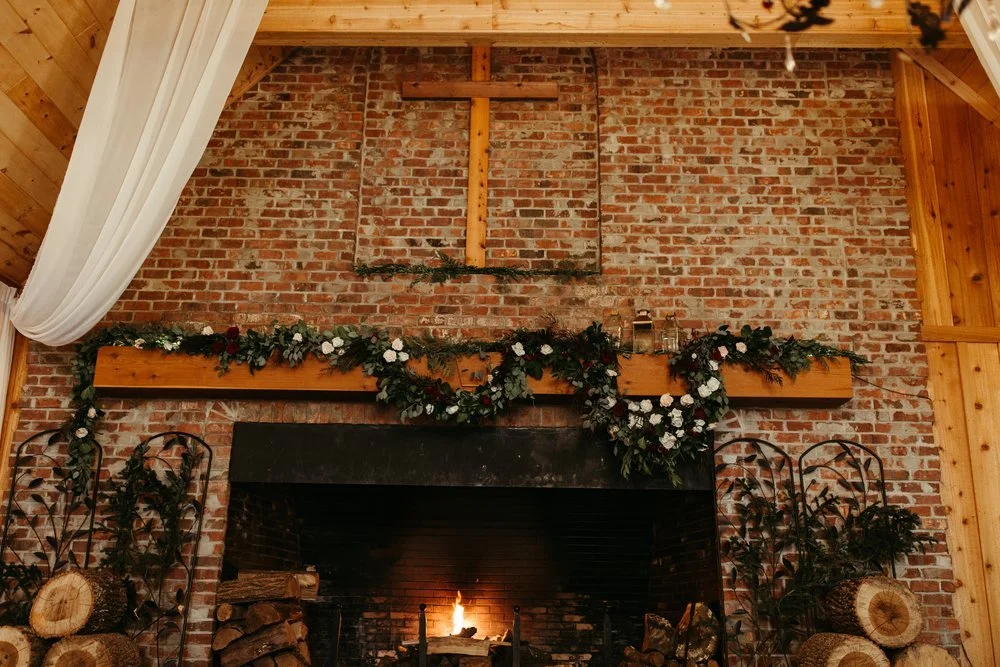 A brick fireplace with a wooden cross above, decorated with flowers and greenery, and logs stacked on either side, suggesting a cozy winter wedding.