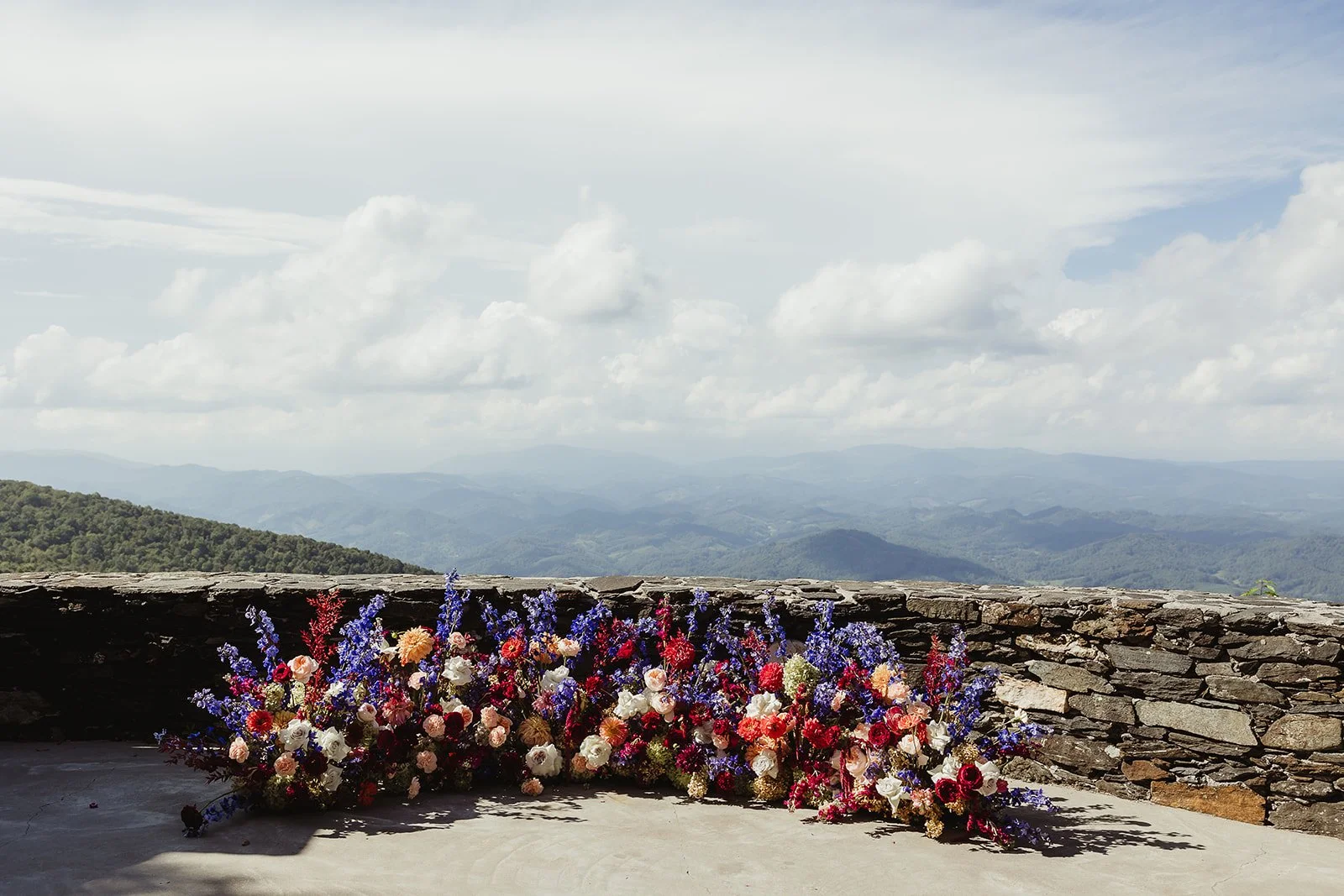 Wide mountain overlook with a long floral arrangement placed along a stone wall under a cloudy sky.