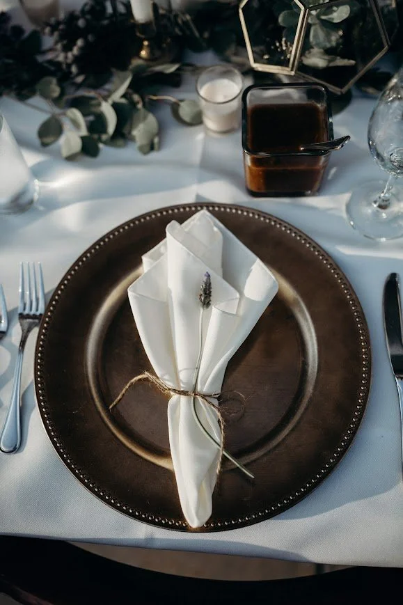 Elegant place setting with a dark charger, folded white napkin tied with twine, and a ring tucked into the napkin fold.