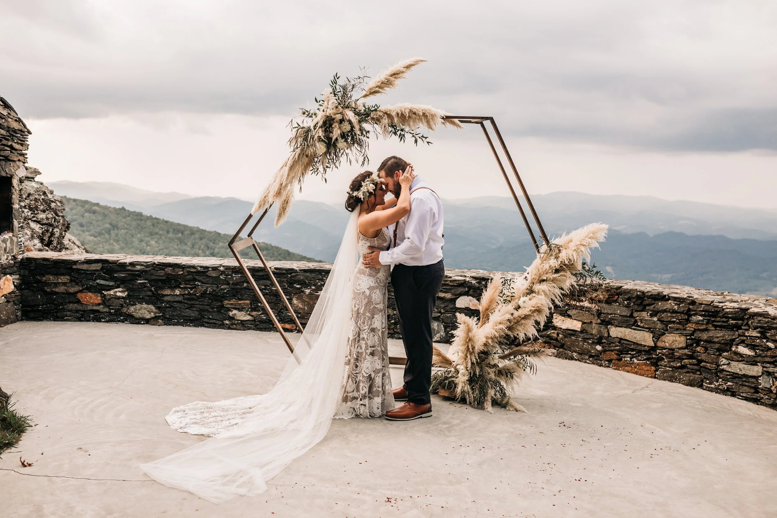 Bride and groom kissing at a mountain overlook ceremony under a geometric arch decorated with pampas grass and greenery.