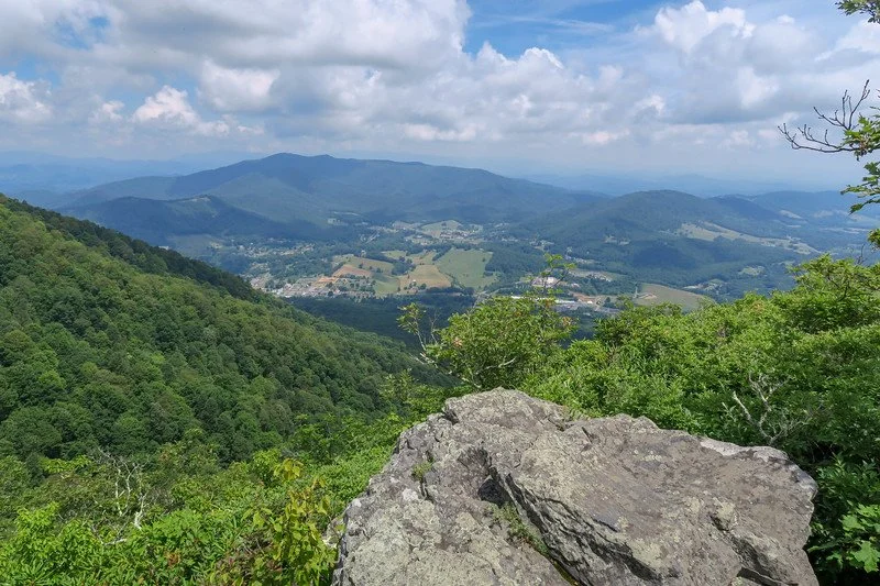 Mountain overlook view across a valley with distant Blue Ridge ridges and large rocks in the foreground.
