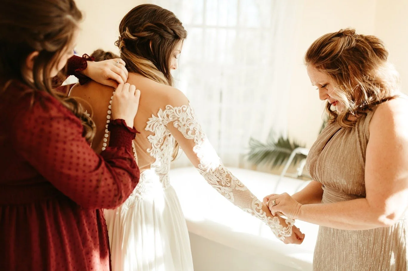 Bridesmaids buttoning the back of the bride’s lace wedding dress in warm window light.