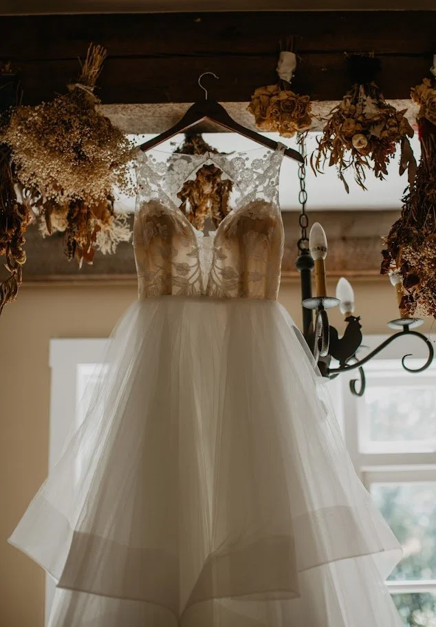 Lace wedding dress hanging indoors beneath dried floral decor and a chandelier before the ceremony.