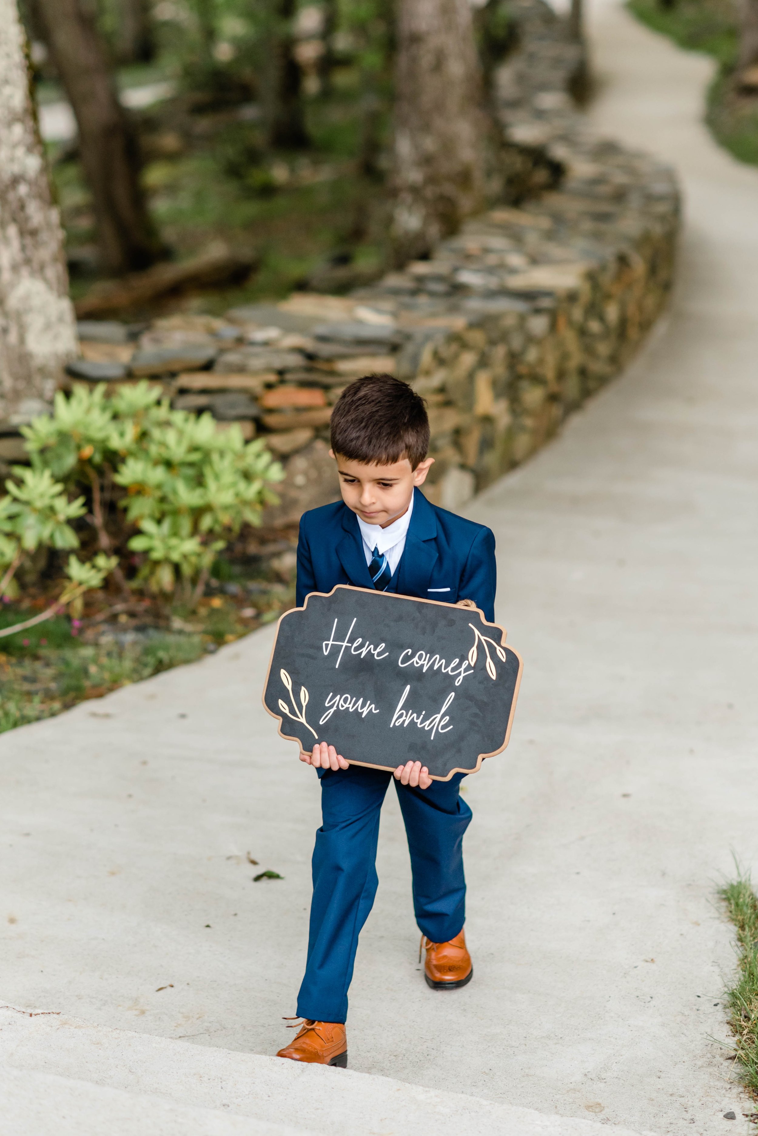 Young ring bearer in a blue suit walking with a sign that says here comes your bride.