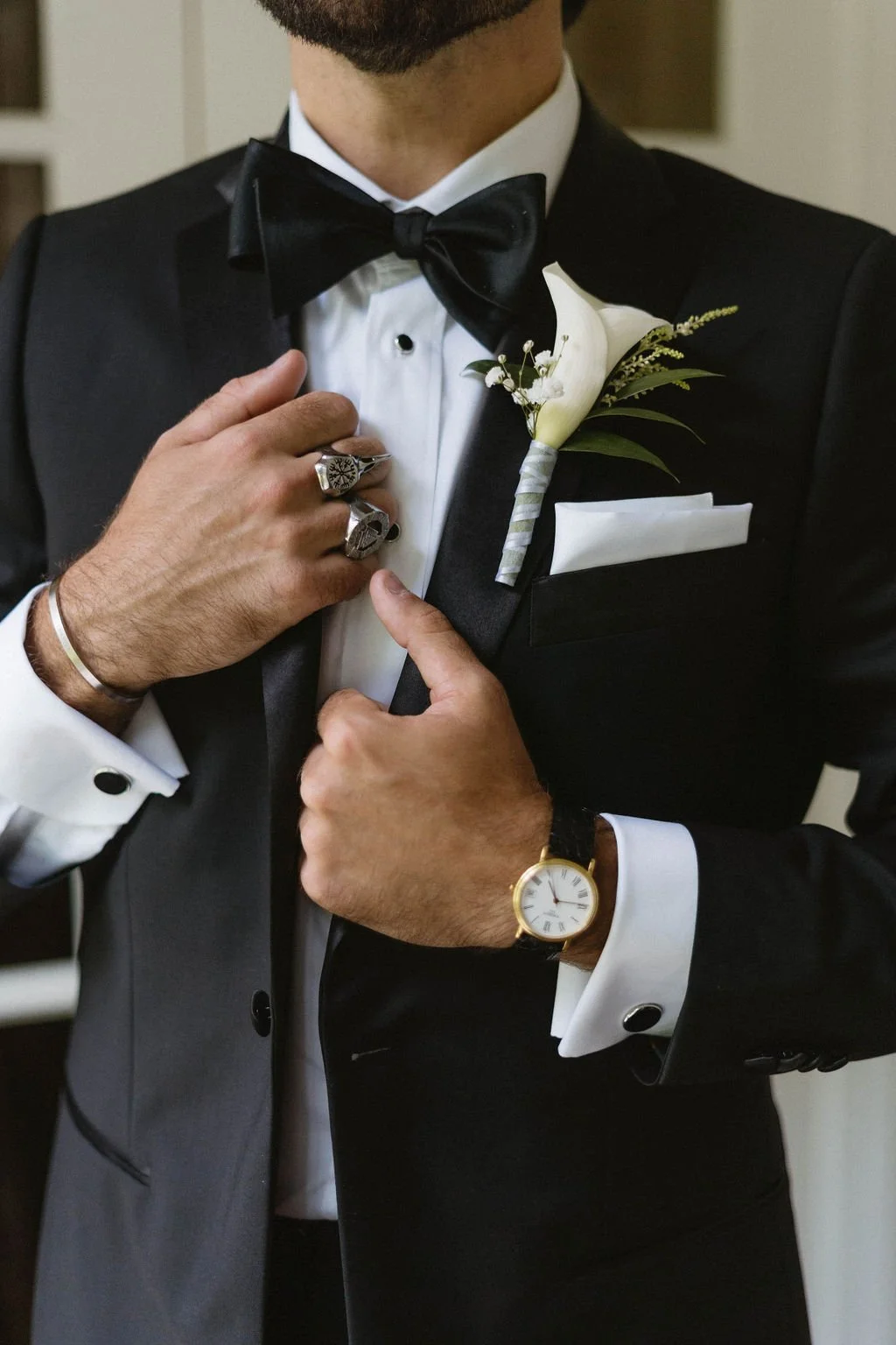 Close-up of a groom in a black tuxedo adjusting his lapel, wearing a calla lily boutonniere and bow tie.