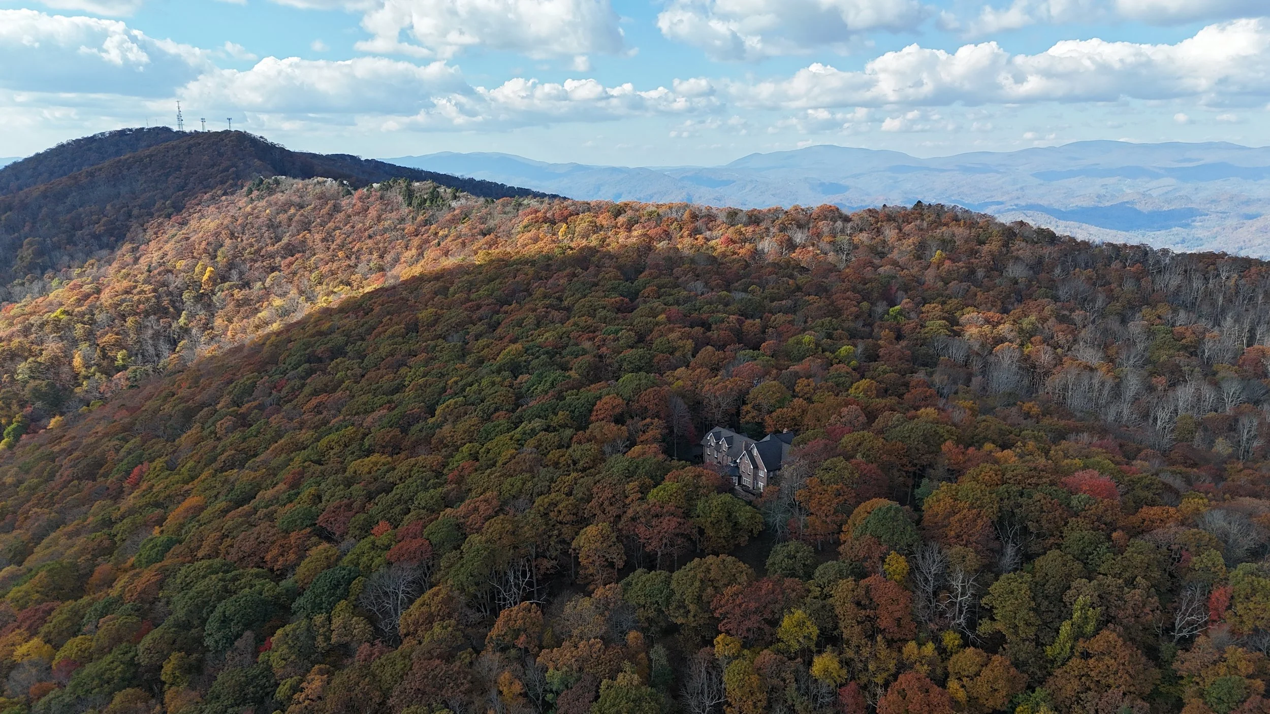 Aerial view of a large brick manor nestled in dense autumn forest, with rolling mountain ridgelines in the distance.