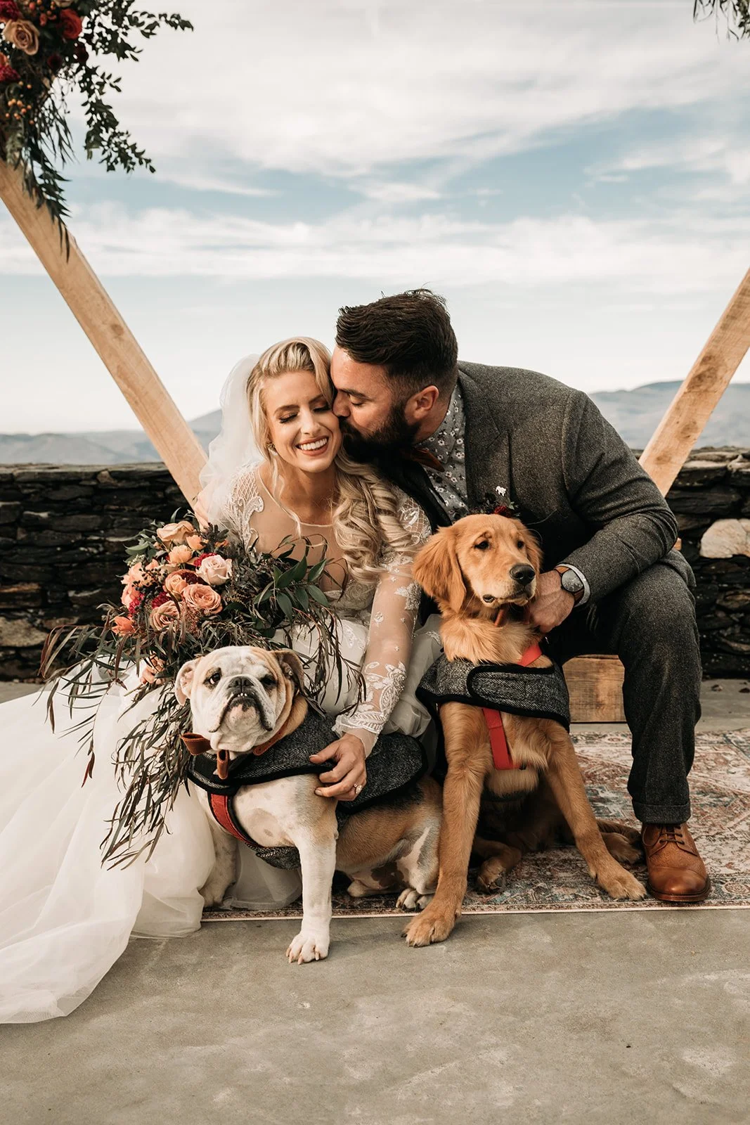 Newlyweds cuddling with two dogs at their mountain wedding ceremony beneath a wooden arch.