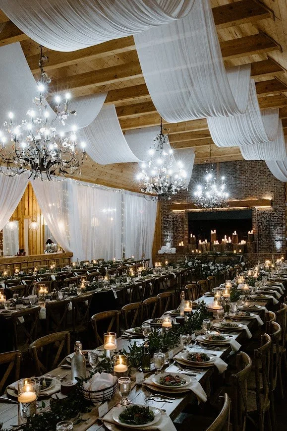 Long reception tables set with black napkins and white candles under warm chandelier lighting.