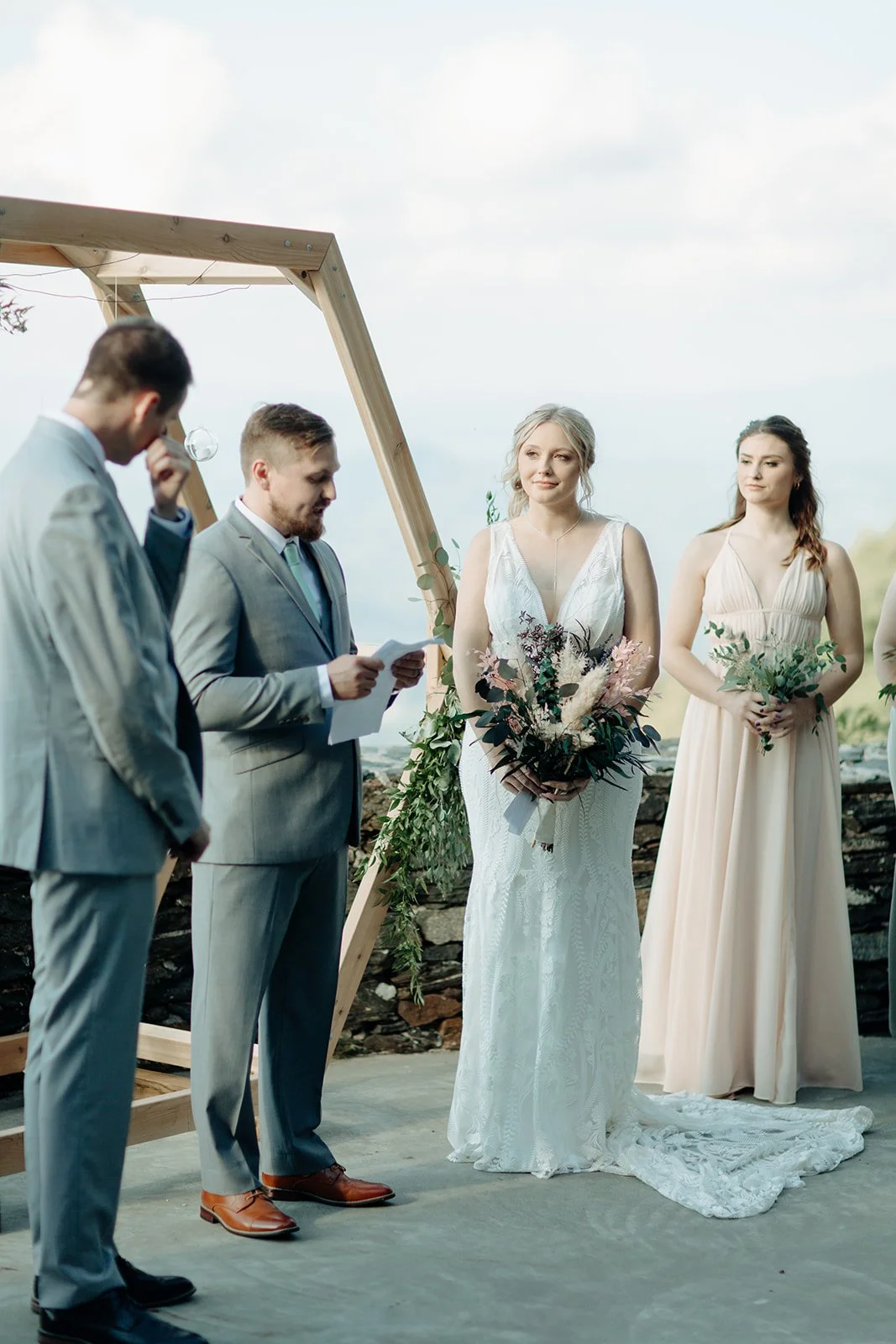 Groom reading vows beside the bride during a mountain ceremony with a hexagon wood arch and pastel bridesmaids.