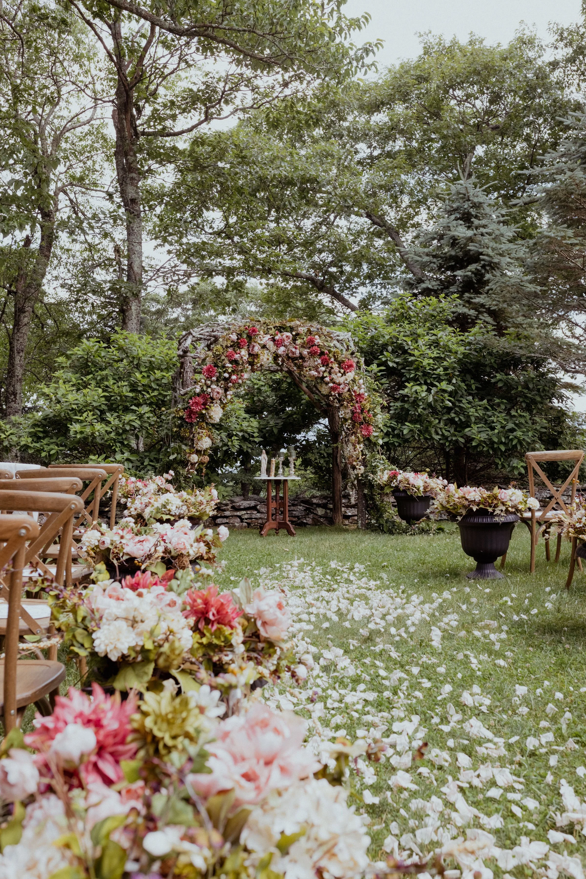 A flower-covered wedding arch and white petals lining a grass aisle in a lush garden.