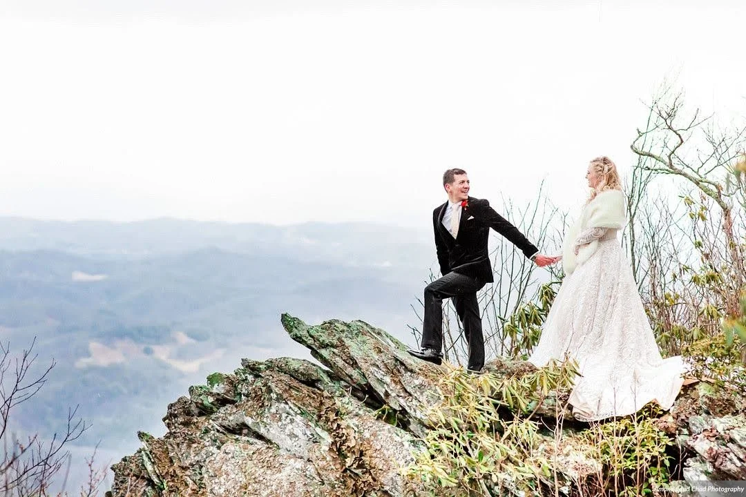 Bride and groom holding hands on a rocky cliff with a mountain view in the background.