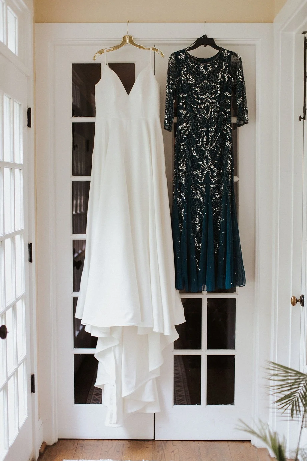 Bride’s white wedding dress and a dark sequined gown hanging on glass French doors.