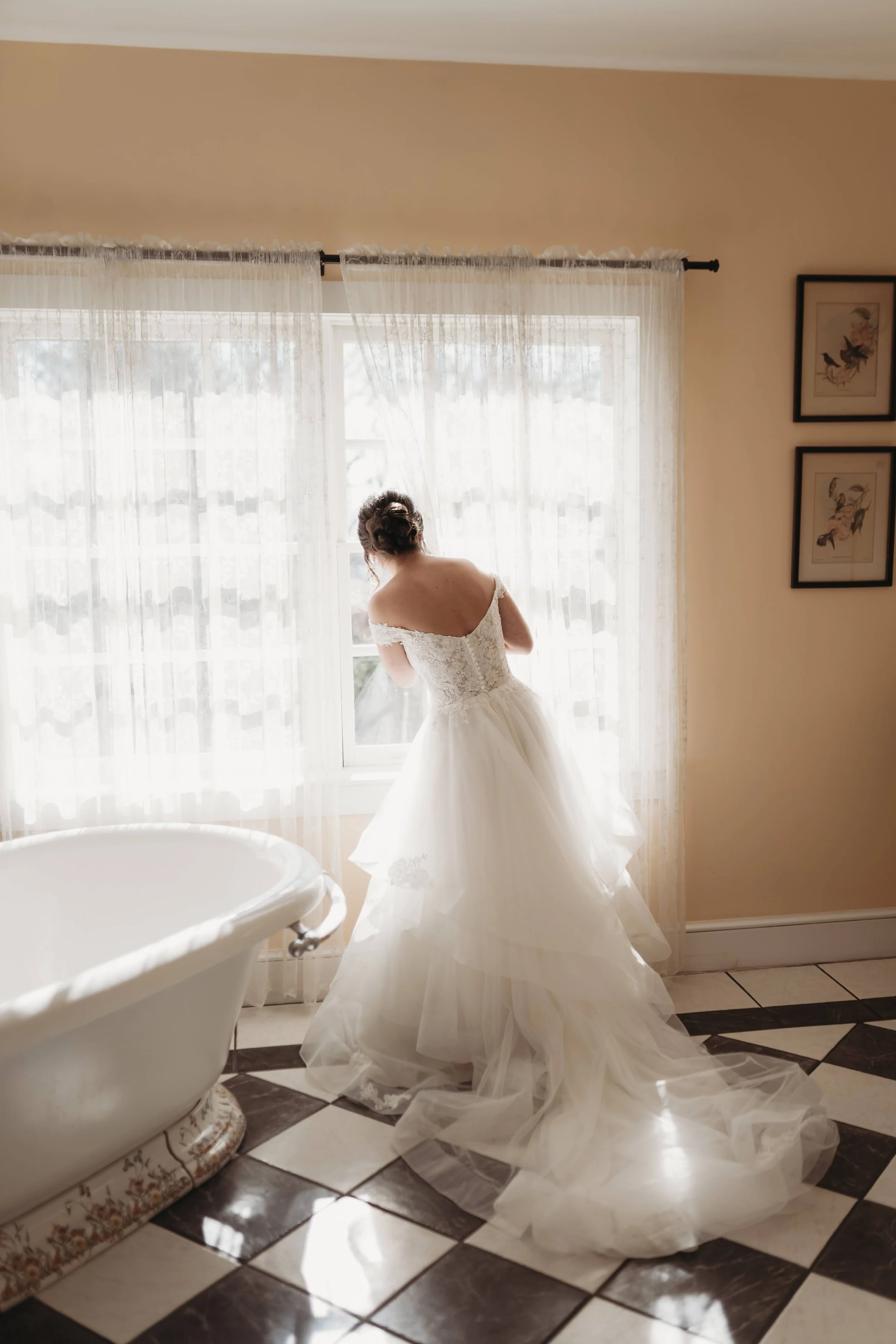 Bride in her wedding dress standing by a bright bathroom window with a vintage tub nearby.
