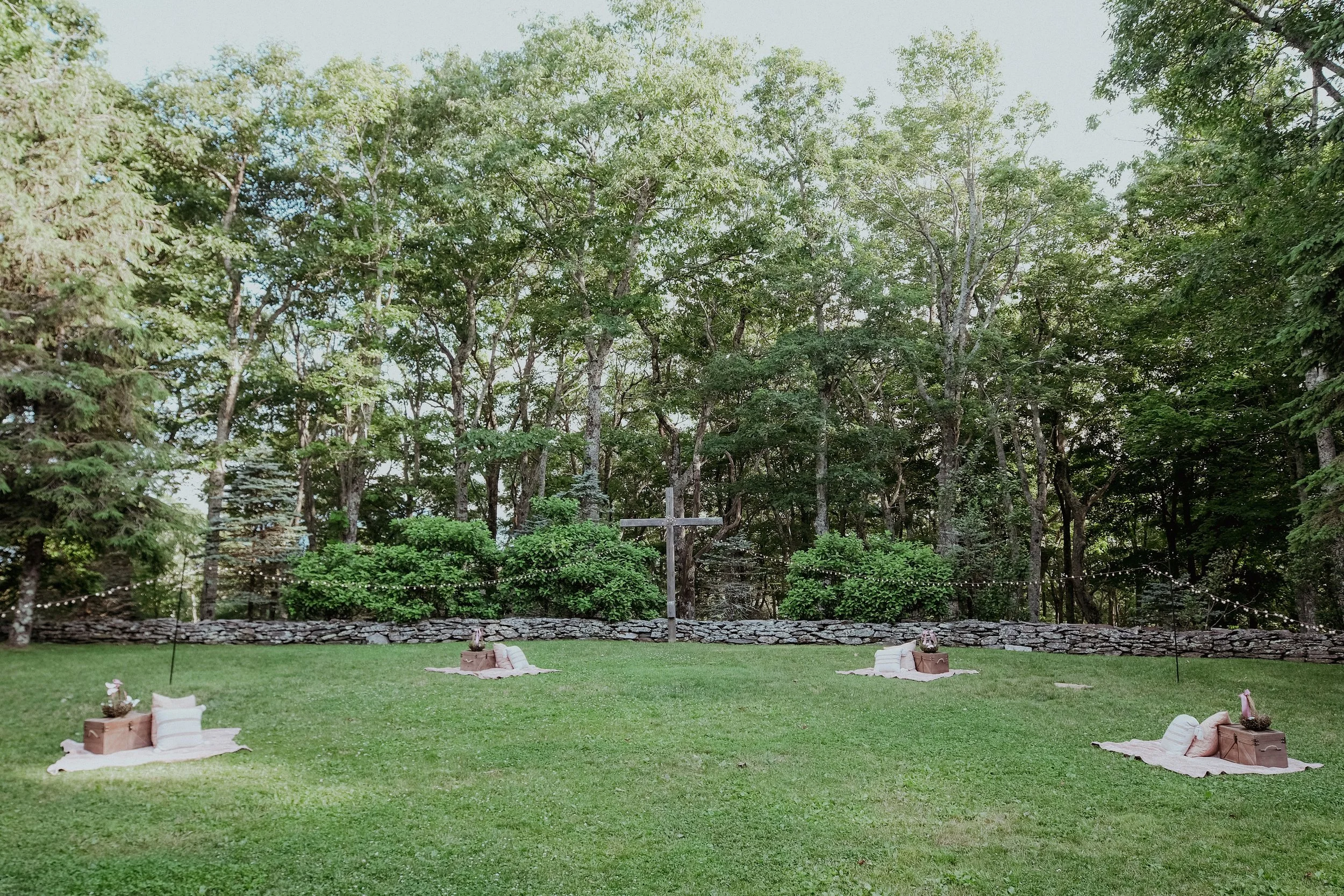 Outdoor lawn ceremony setup with a wooden cross, picnic blankets, stone wall, and wooded backdrop.