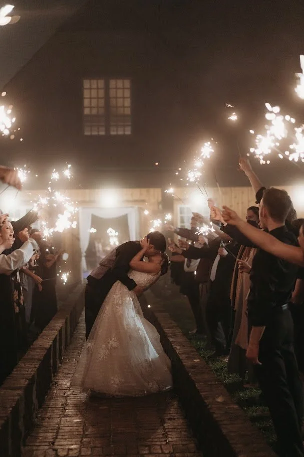Newlyweds sharing a dip kiss during a sparkler exit outside a brick estate wedding venue at night.
