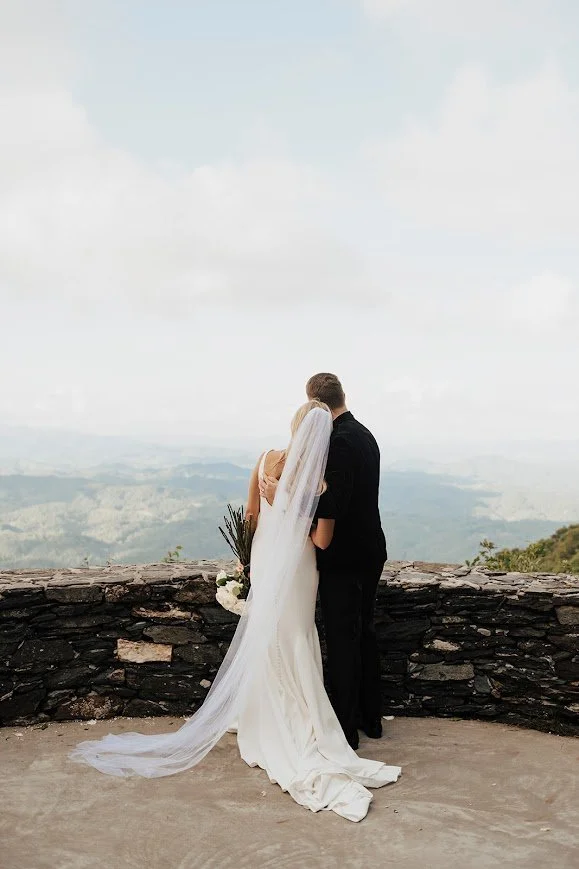Bride in a white wedding dress and long veil embracing her groom on a stone overlook with a panoramic view of lush green mountains.