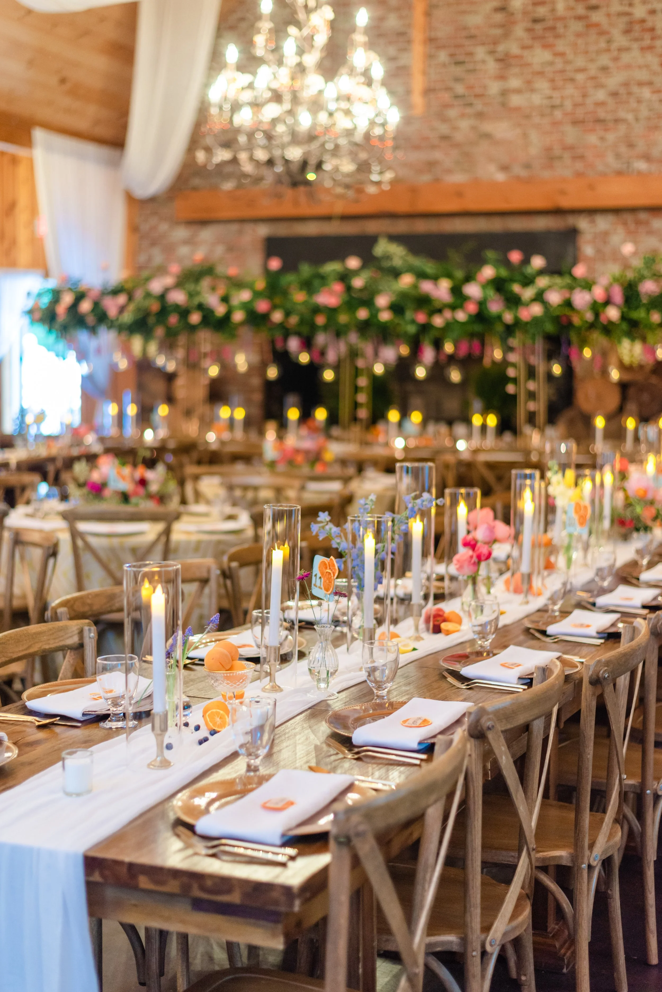 Rustic wedding reception with long tables, colorful flowers, candles, white linens, and chandeliers.