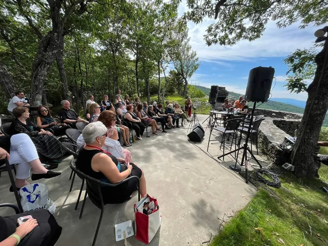 An outdoor event with an audience seated in chairs, listening to speakers on a small stage with a scenic mountain vista in the background under a partly cloudy sky. The setting is surrounded by trees.