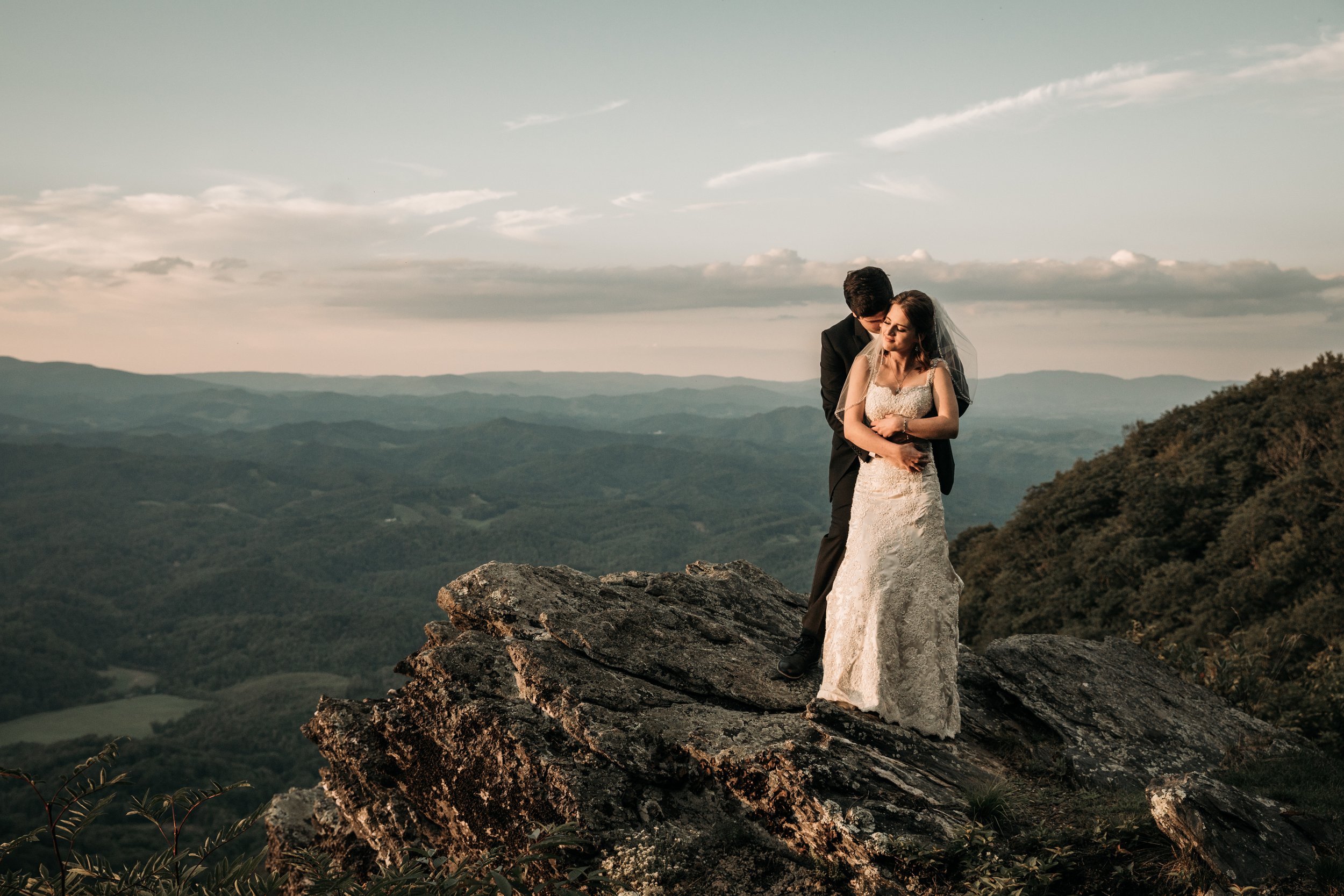 Bride and groom embracing on a rocky mountain overlook with layered mountain views at sunset.