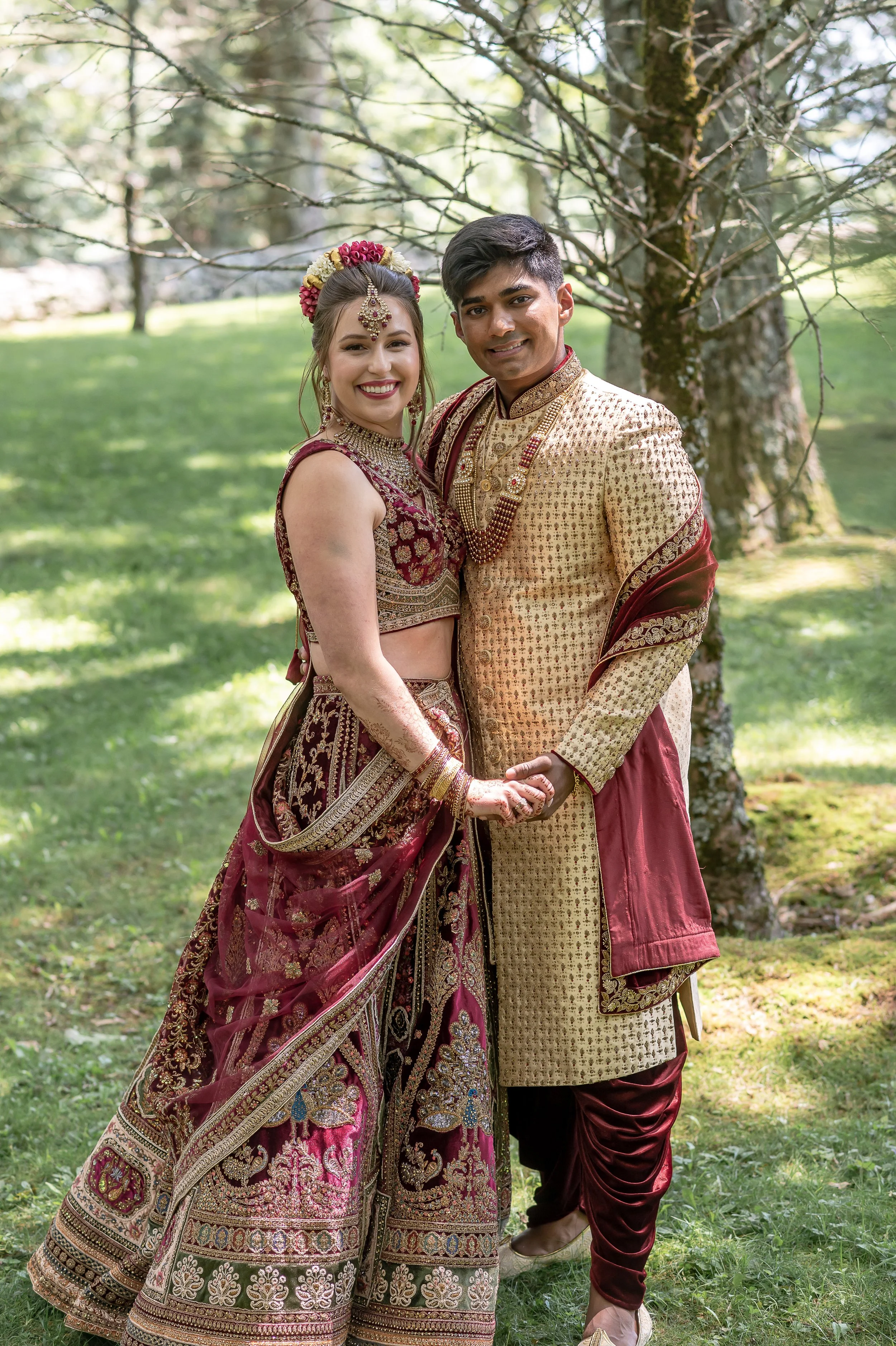 Couple in traditional South Asian wedding attire stands together outdoors among tall trees for a wedding portrait.