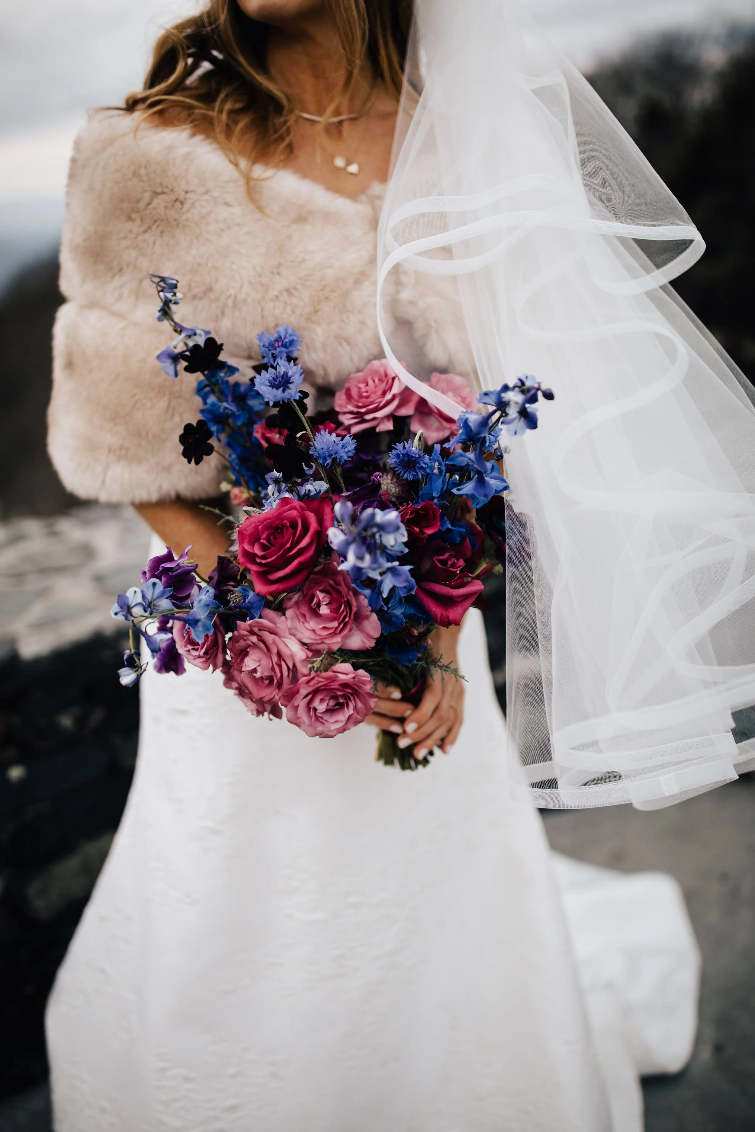 Bride wearing a soft shawl and veil holding a bouquet of pink and purple flowers outdoors.