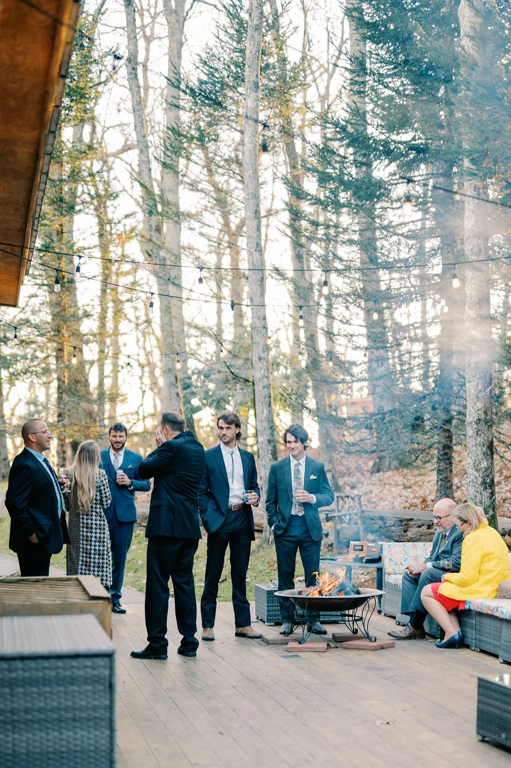 Guests enjoying cocktail hour on a wooded deck with a fire pit, lounge seating, string lights, and drinks.
