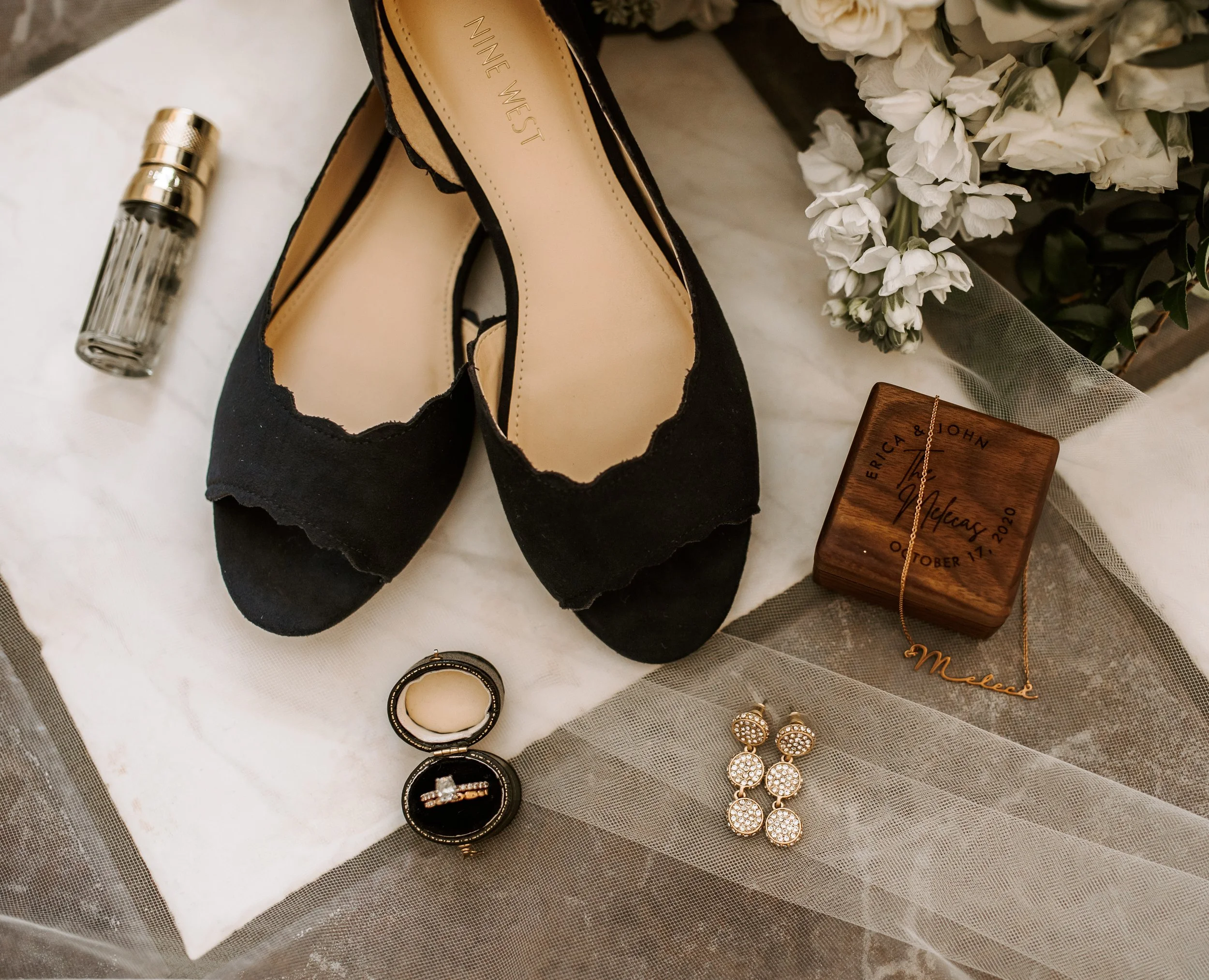 Wedding details flat lay with black heels, rings, perfume bottle, earrings, and a wooden ring box near white flowers