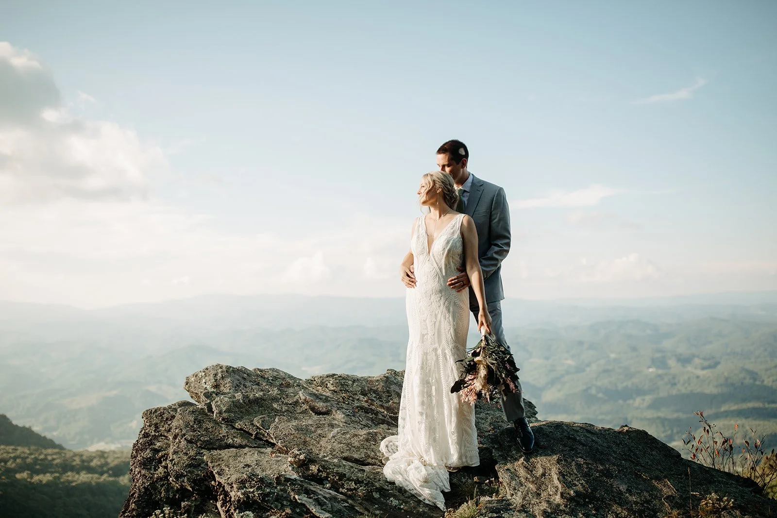 Bride and groom standing together on a rocky Blue Ridge mountain cliff with sweeping valley views behind them.