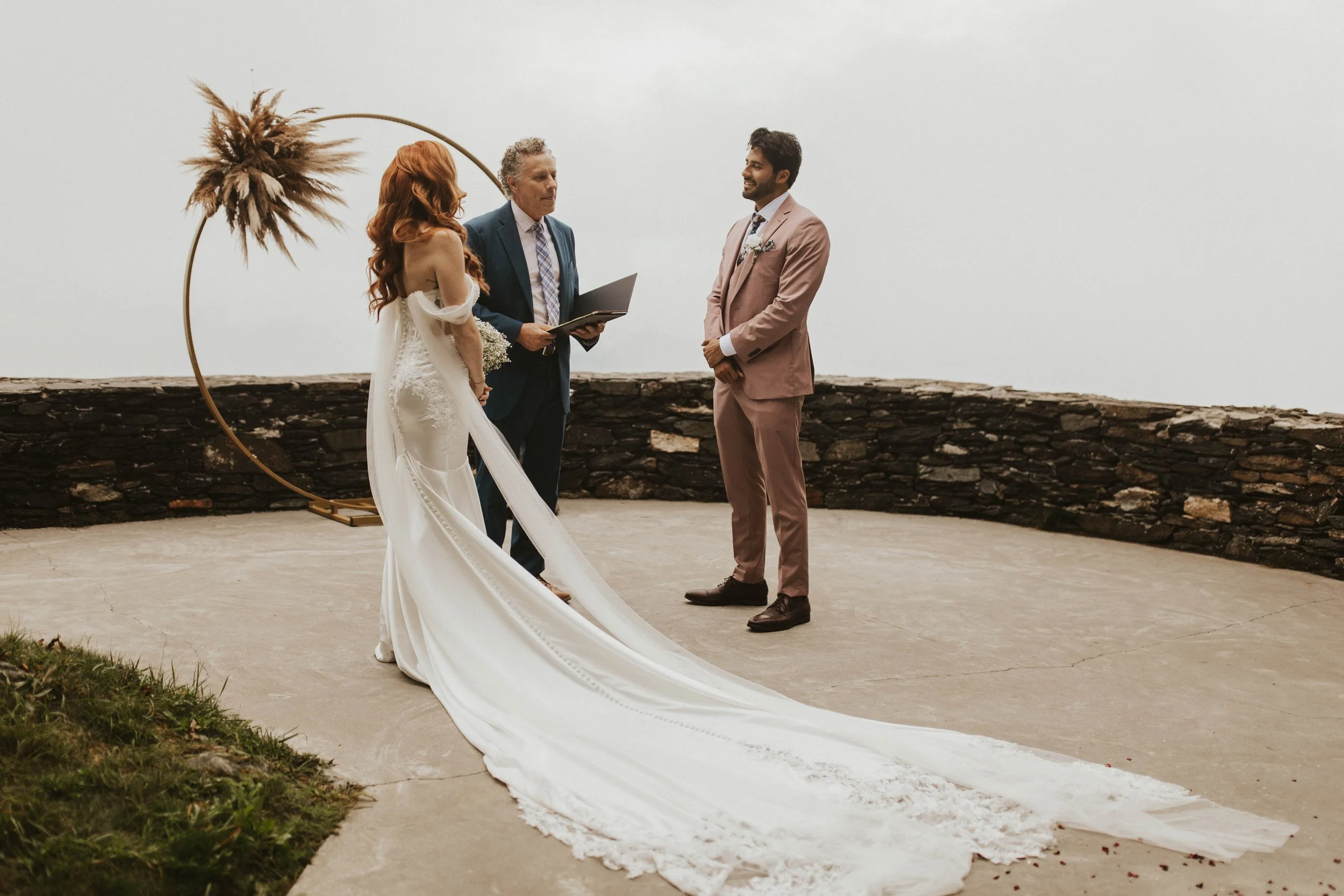 Small mountain overlook ceremony with a circular arch as the bride’s long train trails across the stone patio.