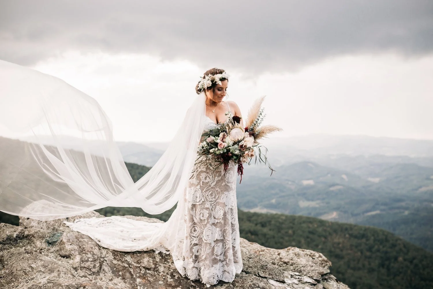 A bride in a white lace dress and floral crown holds a bouquet on a rocky mountain cliff in North Carolina. Her long veil blows in the wind against a backdrop of layered mountains and a cloudy sky.